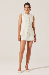 A woman is wearing a sleeveless, cream-colored suit consisting of a fitted, button-front blazer with decorative buttons and a matching mini skirt. She is also wearing cream-colored pointed-toe heels.